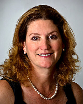 A headshot of a person with wavy hair, wearing a black top and a pearl necklace, smiling slightly.