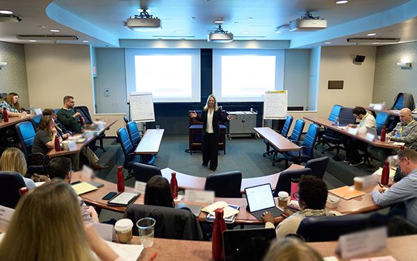 A person giving a presentation in a lecture hall with multiple people seated in tiered seating. There are projectors and screens displaying slides.